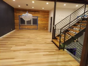 A spacious modern room featuring a newly installed and polished light hardwood floor, a wood plank accent wall, and a metal floating staircase in firestone